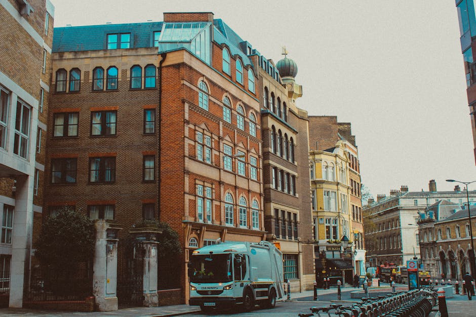 A city street scene featuring a row of multi-story buildings with varied architectural styles and colours. The foreground shows a white waste collection vehicle parked on the pavement, equipped with a